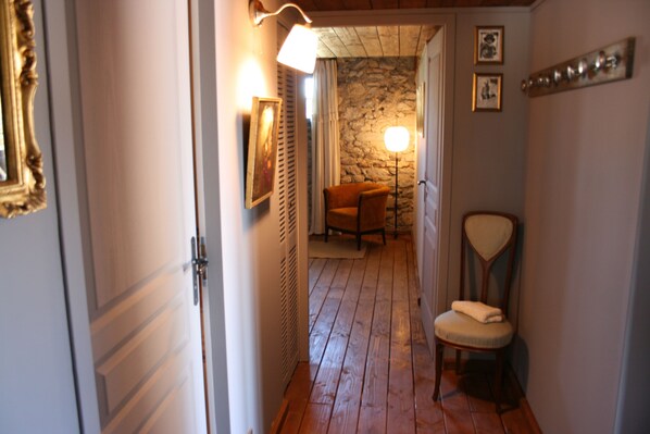 A narrow hallway with wooden flooring, a chair with a cushion, a wall-mounted lamp, and a framed picture.