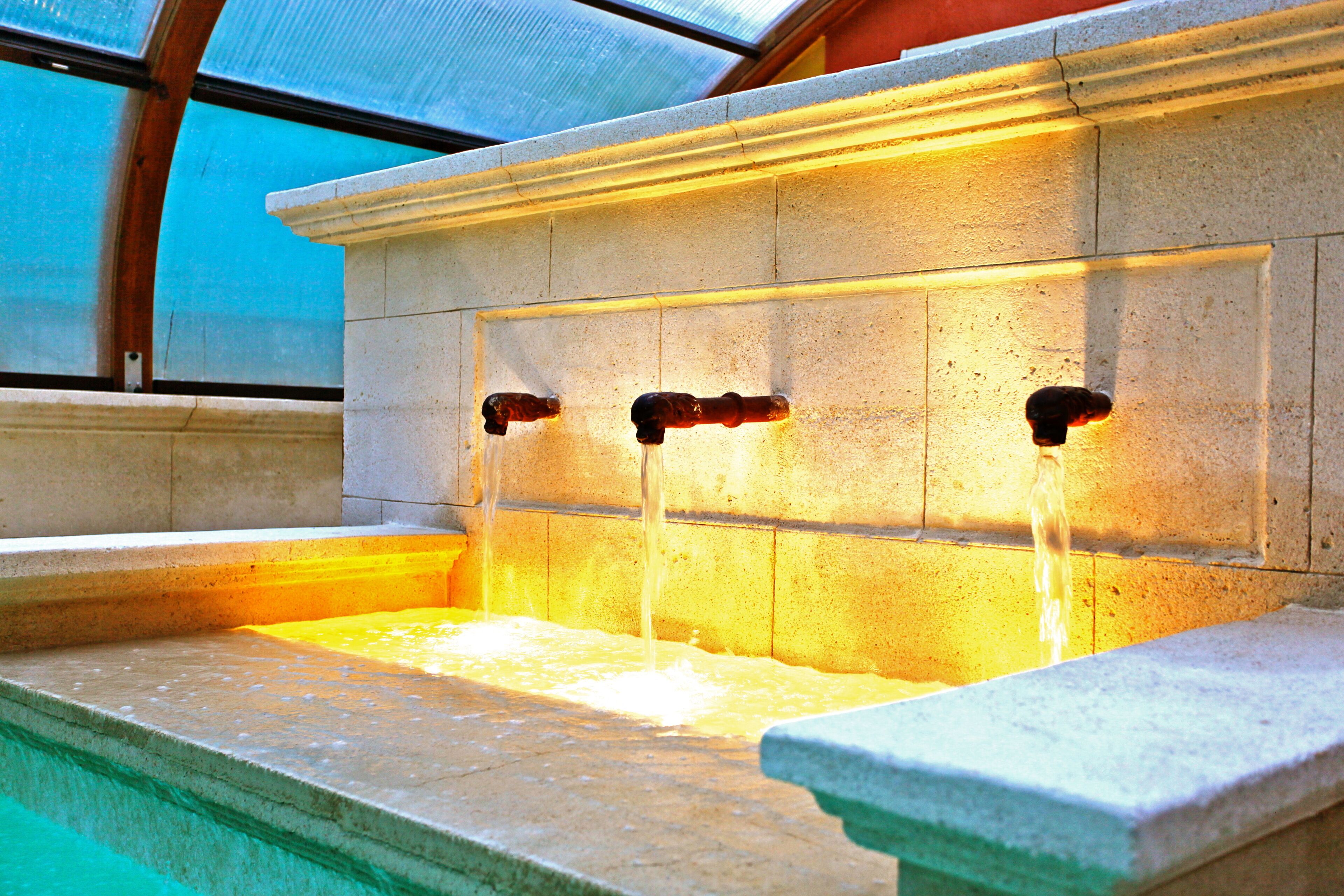 A stone water feature with three spouts, set within a stone structure, likely part of a pool or spa area.