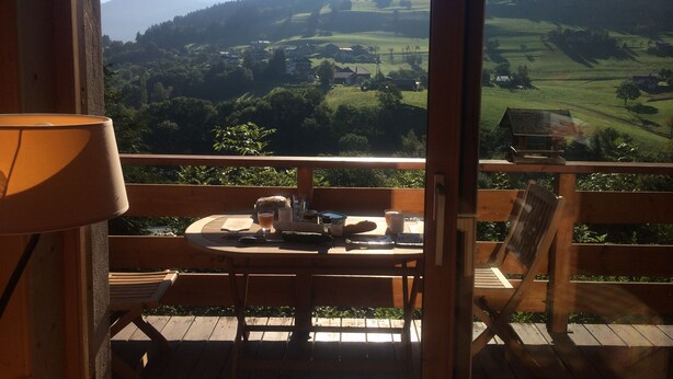 En träveranda med ett bord och stolar, med utsikt över ett bergigt landskap med hus och grönområden.