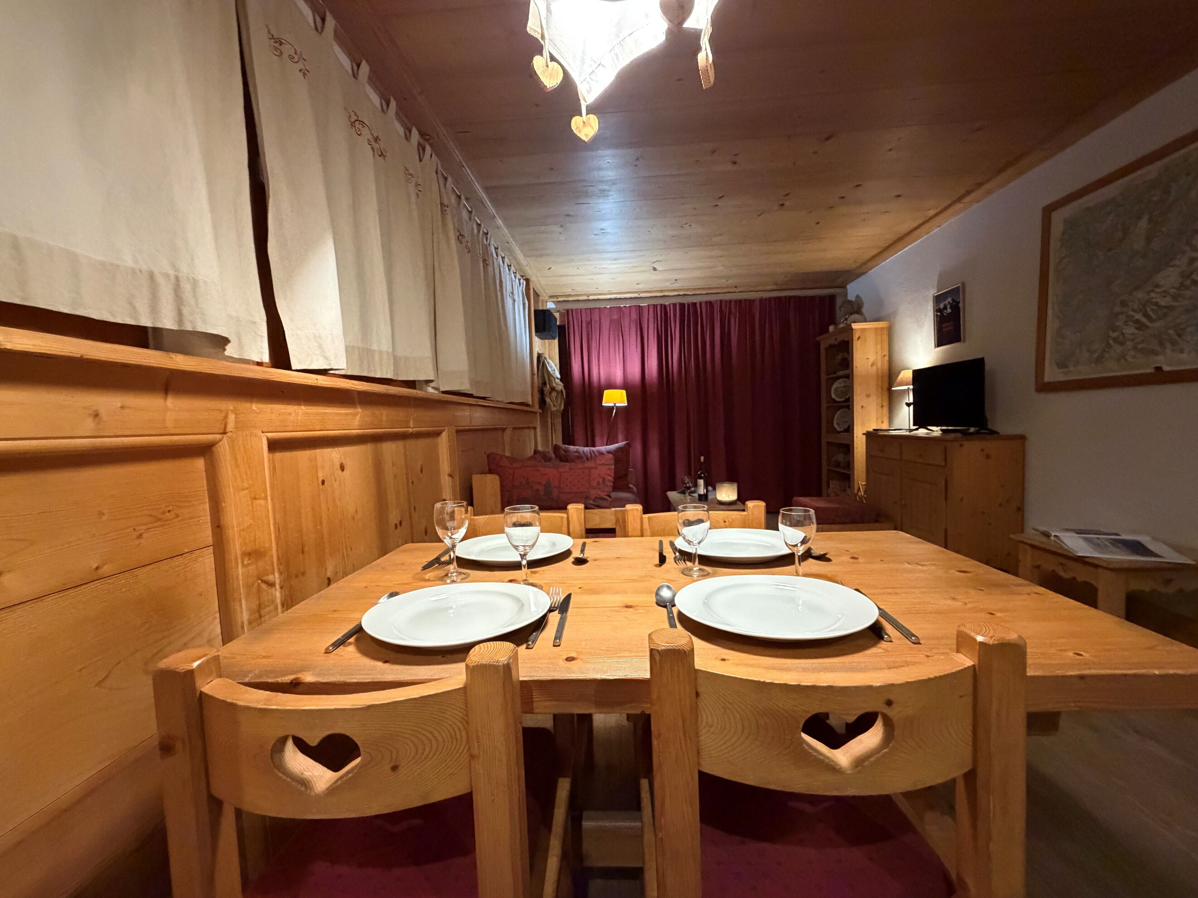 A wooden dining table with four chairs, each with a heart-shaped cutout. The room has wooden paneling and a red curtain in the background.
