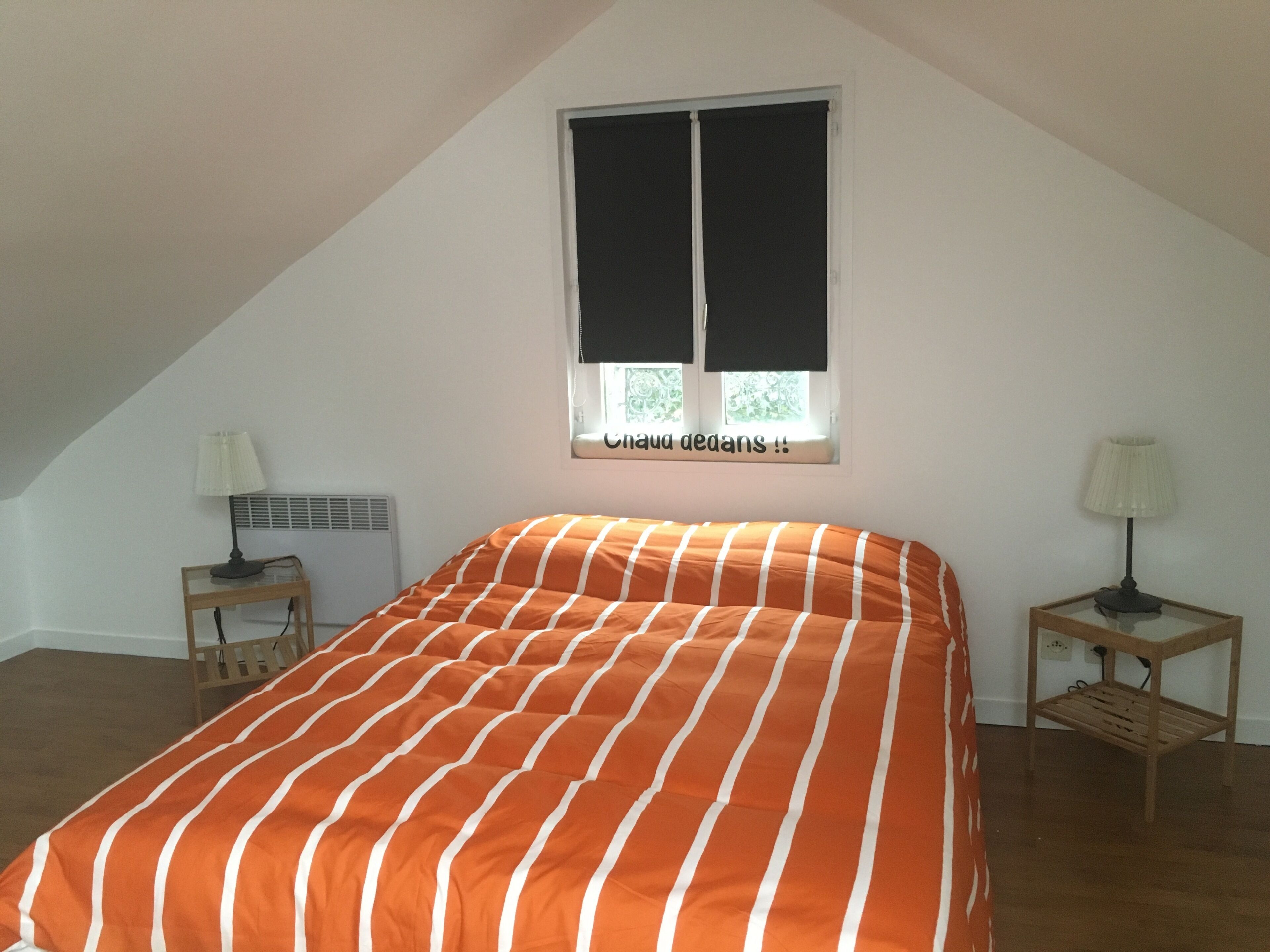 A bedroom with a bed, two nightstands, a window with blinds, and a radiator.