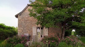 Una casa de piedra con puerta y contraventanas de madera, rodeada de abundante vegetación y plantas en flor.