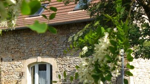 Un edificio de piedra con una ventana y una puerta, rodeado de follaje y flores blancas.