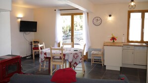 Una sala de estar acogedora con un sofá, una butaca roja, una mesa de comedor con sillas, una zona de cocina con un fregadero y armarios, y un televisor montado en la pared.