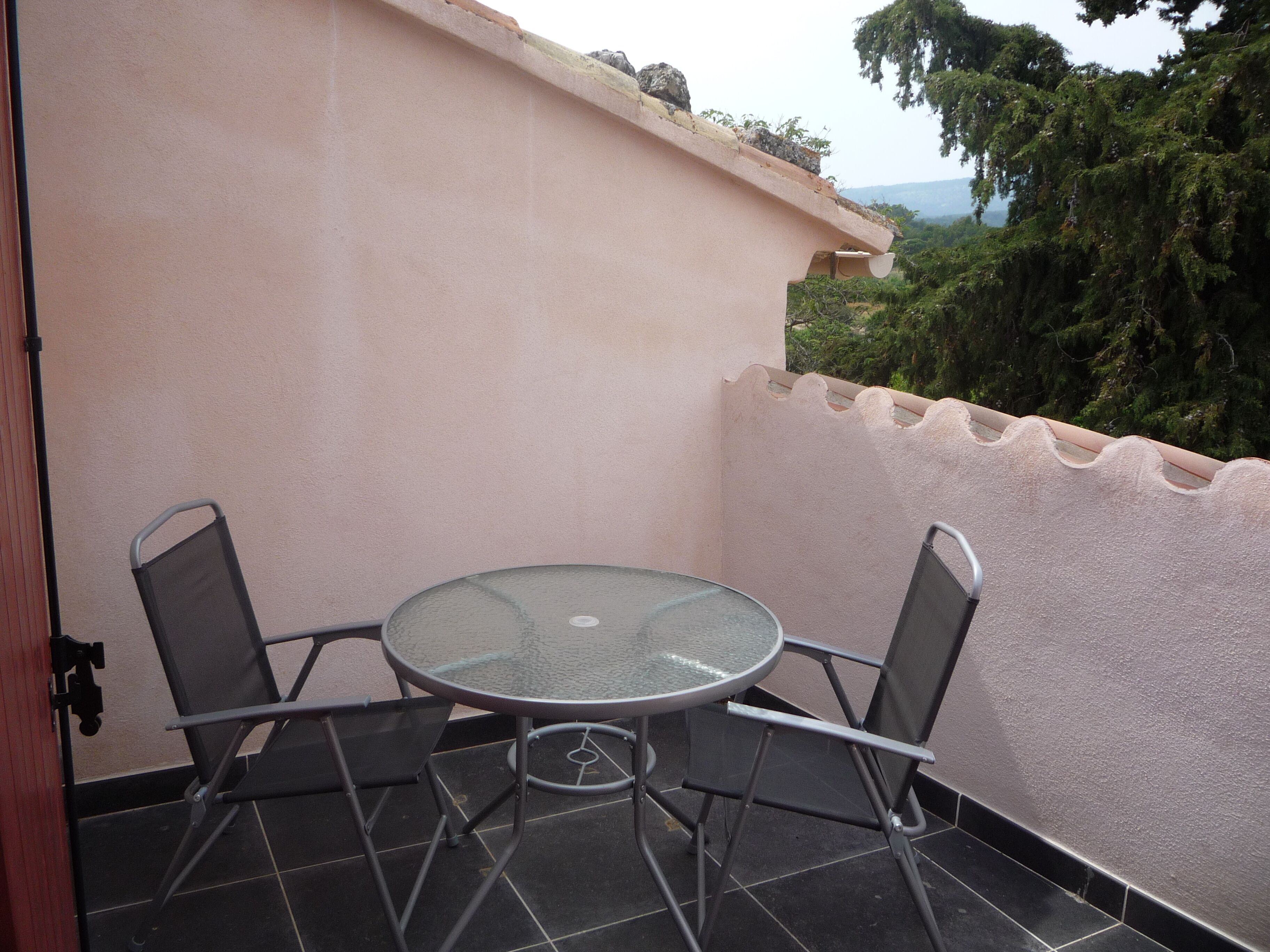 Un petit balcon extérieur avec une table en verre et des chaises en métal, entouré d’un petit muret et de verdure.