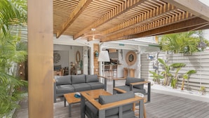 Un patio al aire libre con suelo de madera, sofás grises, mesas de madera y un techo con listones de madera.