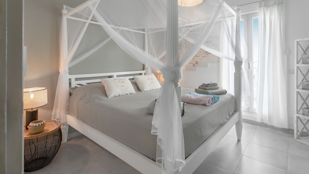 Una cama con dosel y cuatro columnas con acabado blanco, una mesita de noche con lámpara, y vista al exterior a través de una ventana con cortinas traslúcidas.
