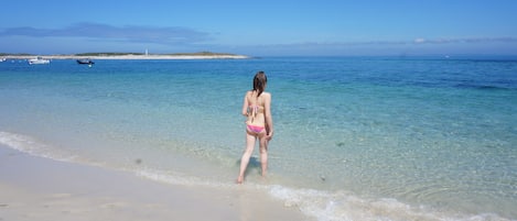 Una playa con agua cristalina, una persona en traje de baño y barcos a lo lejos.