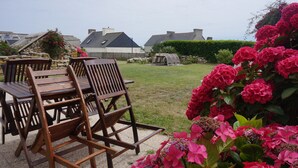 Une terrasse avec des chaises et une table en bois, entourée d’un jardin où poussent des fleurs roses et bordée d’un mur de pierre.