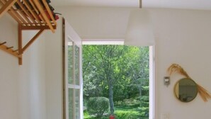 Una habitación con un aparador de madera, un armario blanco, un espejo en la pared y vistas a un jardín verde a través de una puerta abierta.