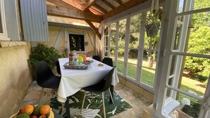 Une terrasse couverte avec une table dressée pour deux personnes, agrémentée d’une nappe blanche, de chaises et d’un plateau contenant de la nourriture et des boissons.