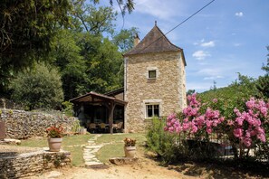 A stone building with a tower, surrounded by trees and a garden with pink flowers. - House rental w/pool, Jacuzzi, sauna in the Périgord Noir, 5 km away from Sarlat (Vézac)