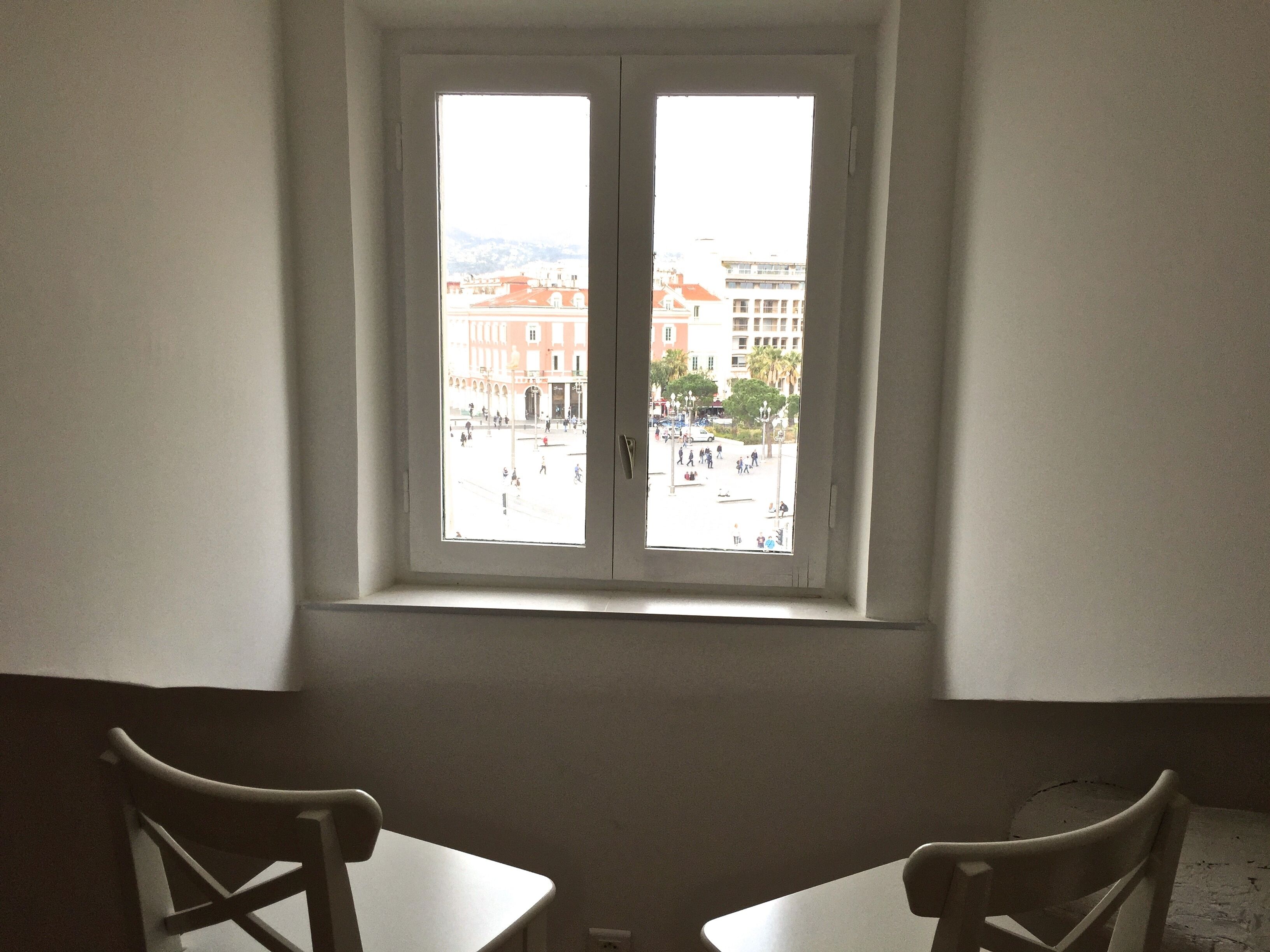 A room with a window overlooking a public square with buildings and people. There are two white chairs and a small table in the foreground.