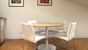 Ein Essbereich mit einem runden Tisch und vier weißen Stühlen, zwei gerahmte Bilder an der Wand und ein Küchenbereich mit Holzboden.