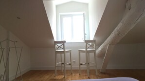 Ein Dachzimmer mit geneigter Decke, zwei weiße Stühle, ein Metallregal und ein Fenster mit Blick nach draußen.