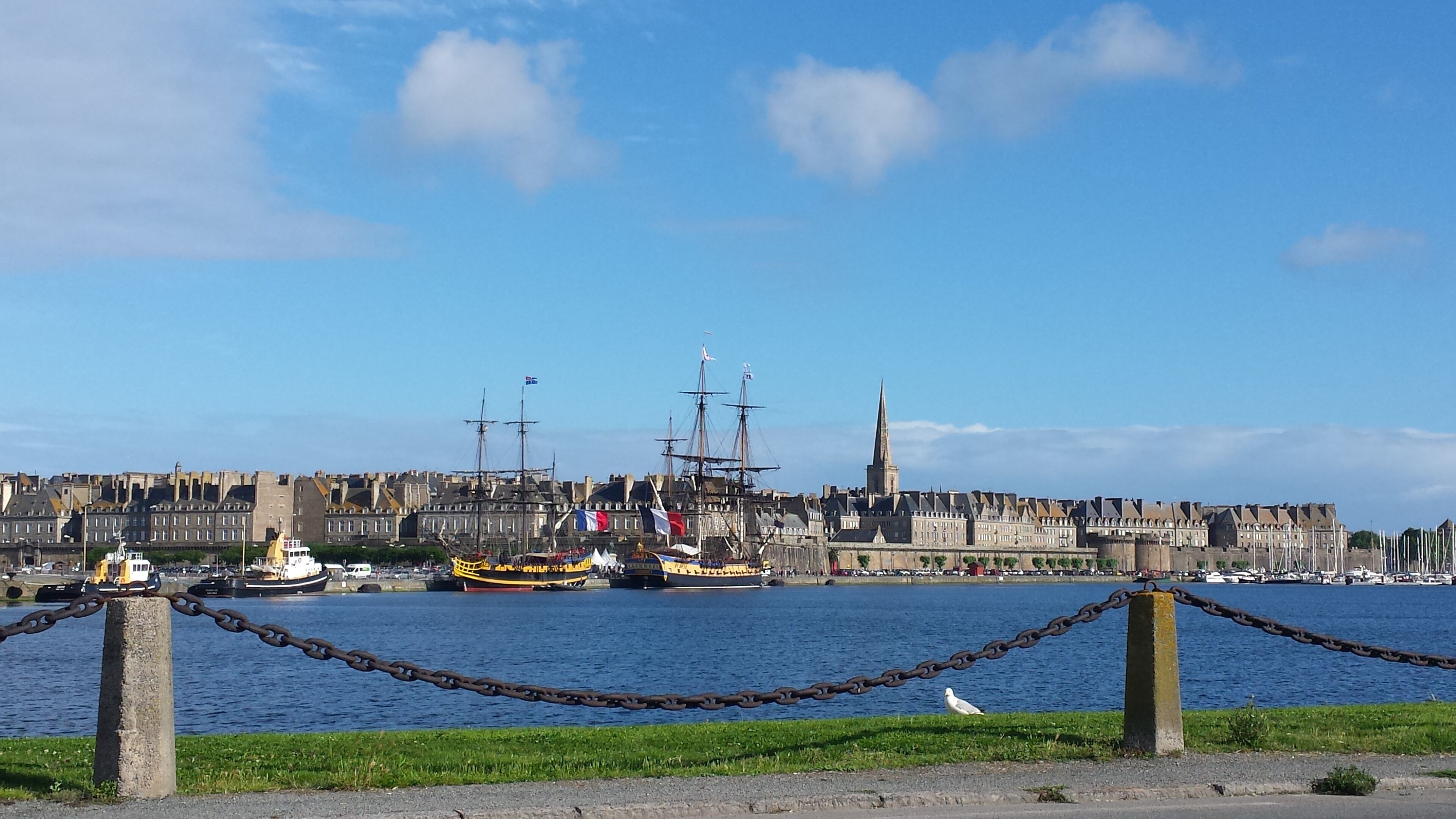 Un paysage urbain au bord de l’eau, avec des bâtiments historiques, un clocher d’église imposant et une variété de bateaux amarrés le long du port.