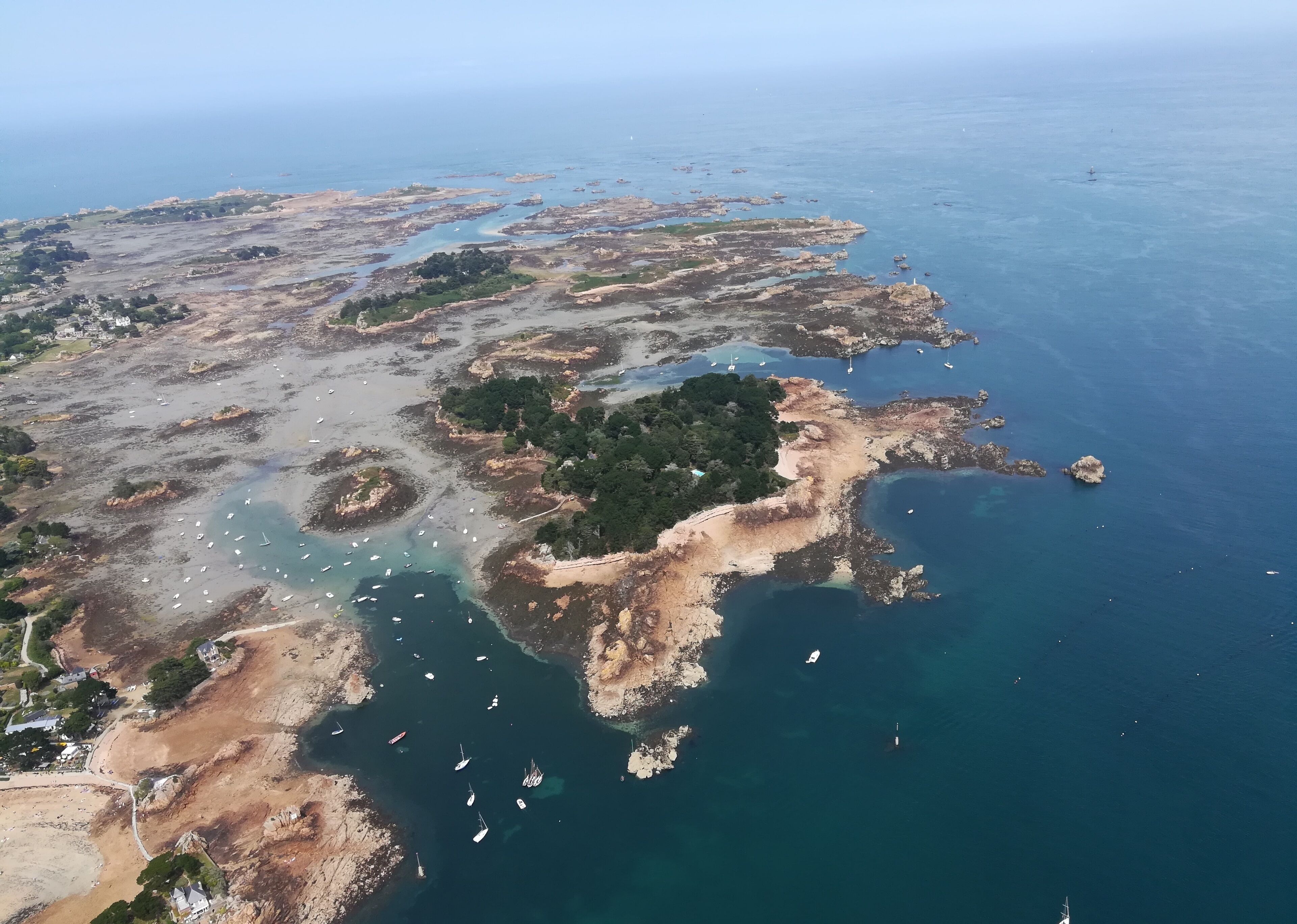 An aerial view of a coastal area with rocky terrain, clear blue waters, and several boats.