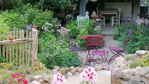 A garden with a wooden fence, a red metal table and chair, and various flowering plants. - A cottage in a garden in Rougegoutte (Rougegoutte)