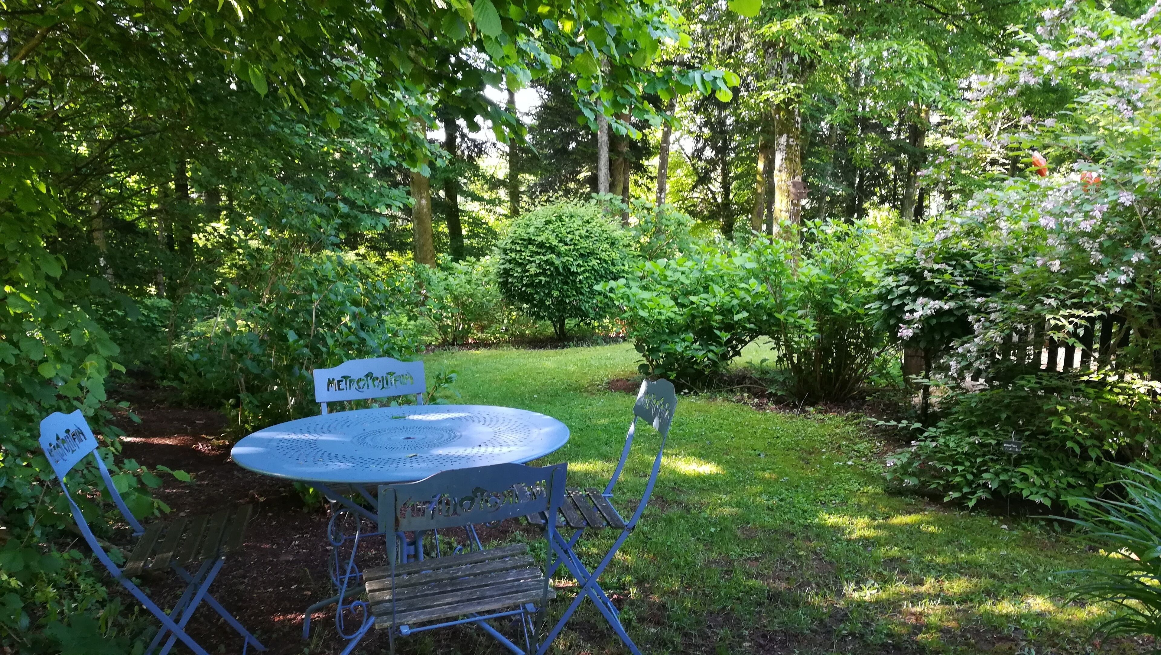 Een ronde metalen tafel met een bord waarop 'MetroRural' staat, omringd door blauwe metalen stoelen in een weelderige tuin.