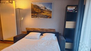 A neatly made bed with white linens in a room with a mountain landscape painting on the wall, a radiator, and a shelf with board games.