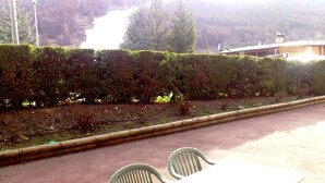 A paved outdoor area with a table and two chairs, a neatly trimmed hedge, and a building in the background.