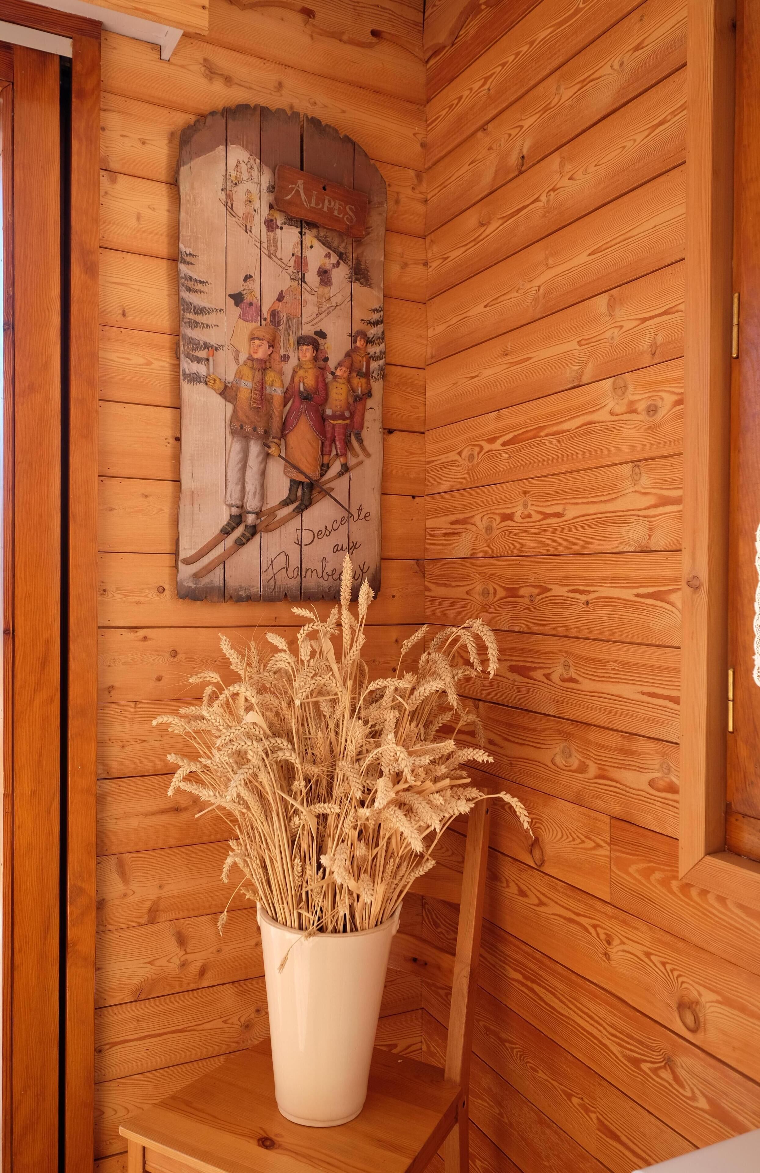Een houten blokhut van binnen, met een houten wand, een lijst met een foto van skiërs en een witte vaas met gedroogde planten.