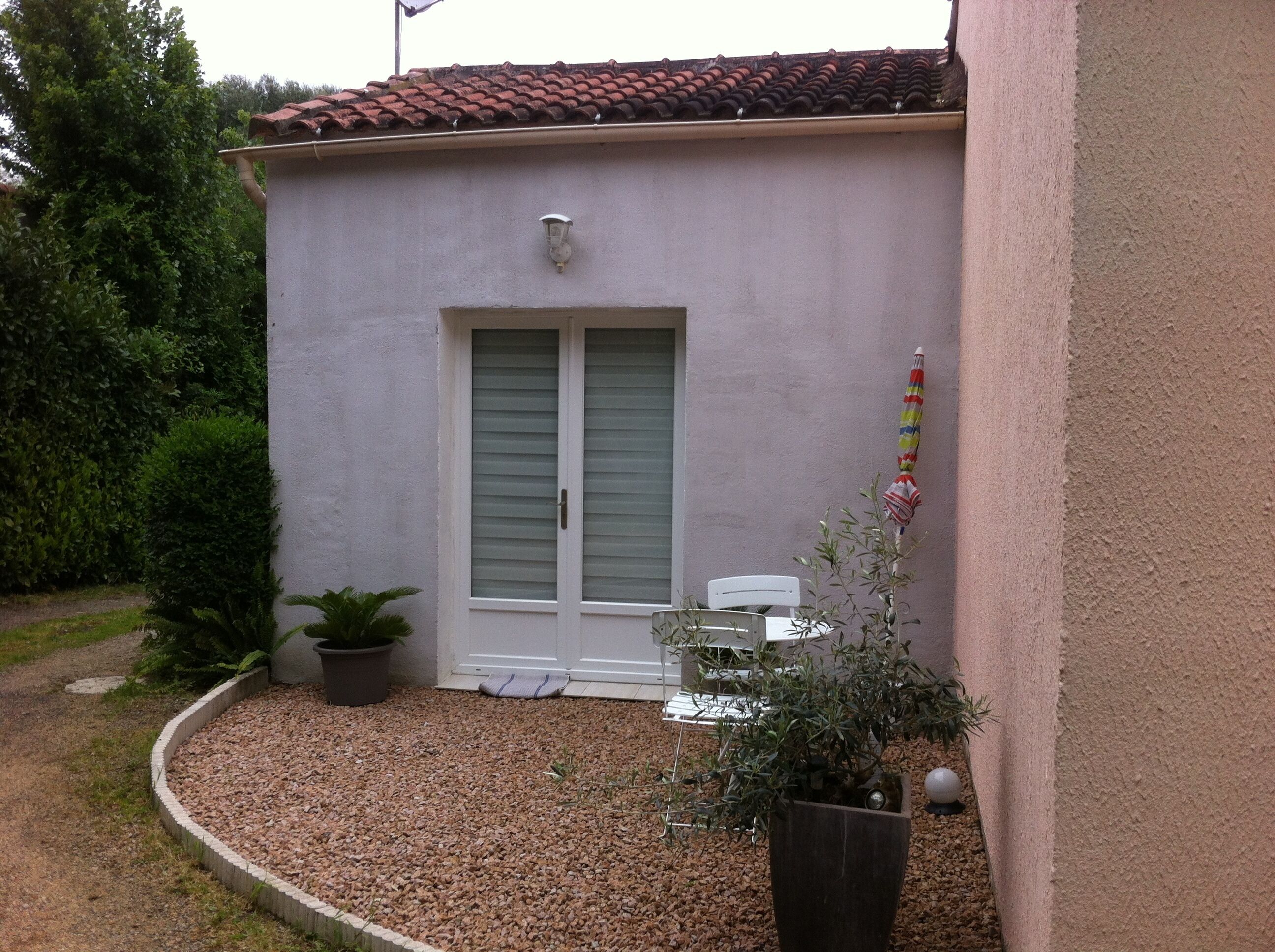 Un petit bâtiment avec un toit en tuiles, une porte avec des volets, un pot de fleurs et une petite table avec une chaise.