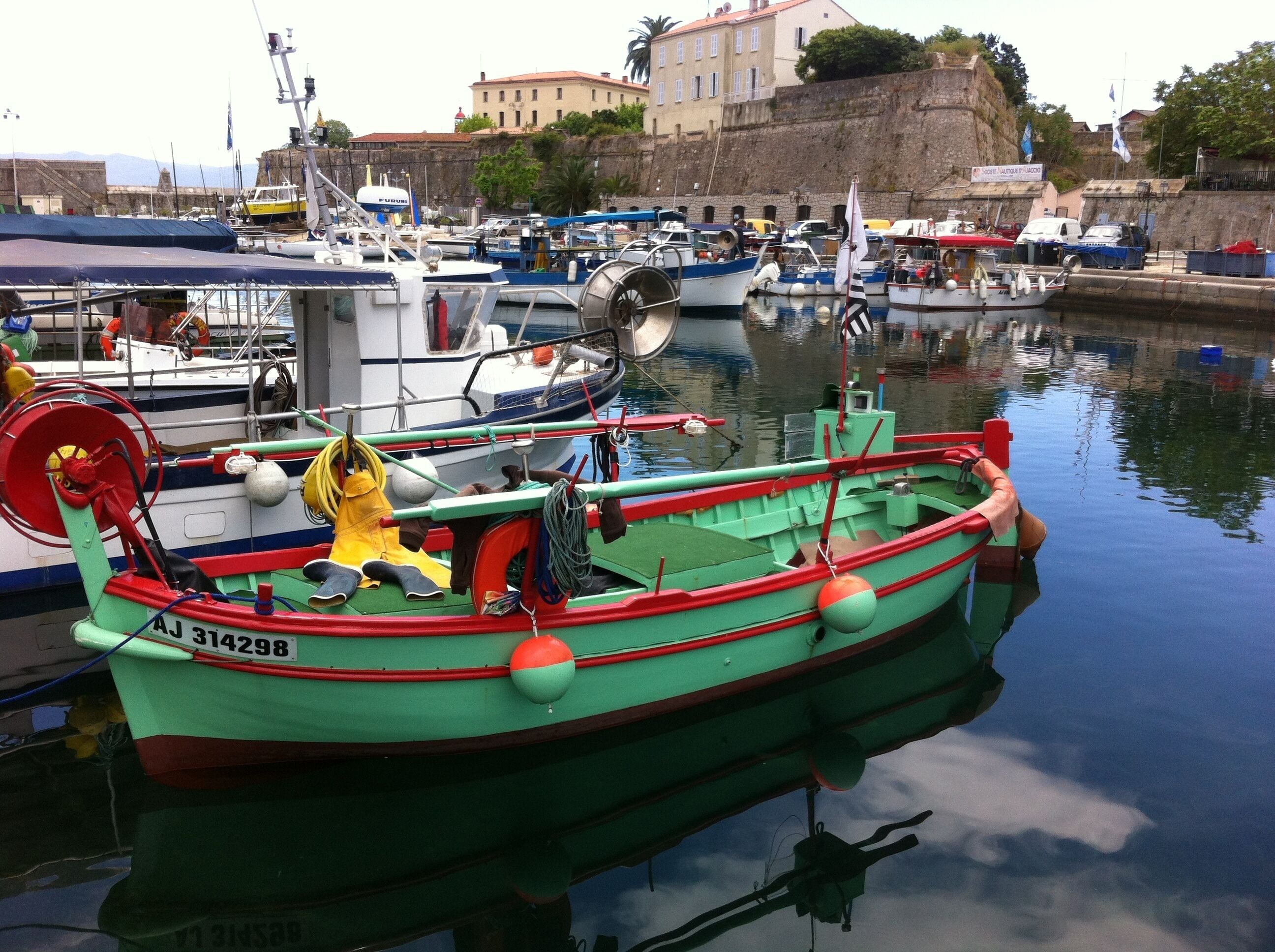 A harbor with various boats docked, a building with a red roof in the background, and a stone wall.