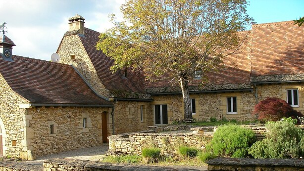 A stone building with a sloped roof, a wooden door, and a small garden area with a tree and shrubs. - Cottage of charm and character, 220 m2, quiet, between Sarlat and Lascaux. (Archignac)
