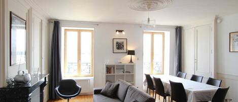 Ein modernes Wohnzimmer mit einer Couch, einem Esstisch und Stühlen. Zwei Fenster mit Vorhängen, eine Deckenleuchte und ein Bücherregal sind vorhanden.