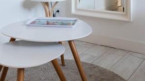 Una habitación moderna con una mesa redonda blanca, una lámpara y una pequeña mesita auxiliar blanca. Un espejo grande refleja una puerta y un televisor.