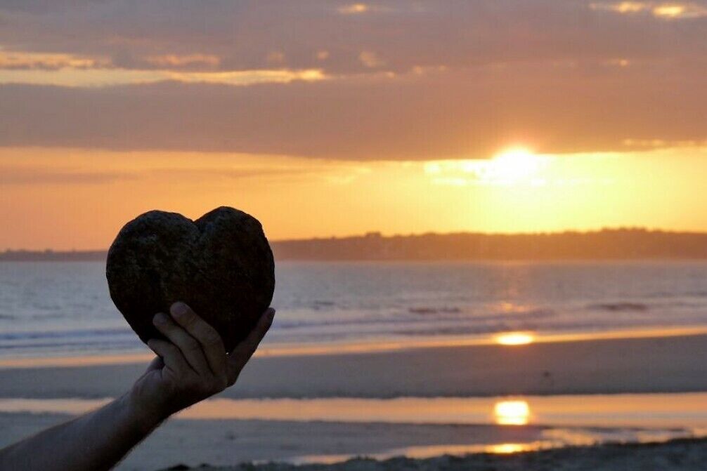 A hand holding a heart-shaped rock against a sunset over a body of water.