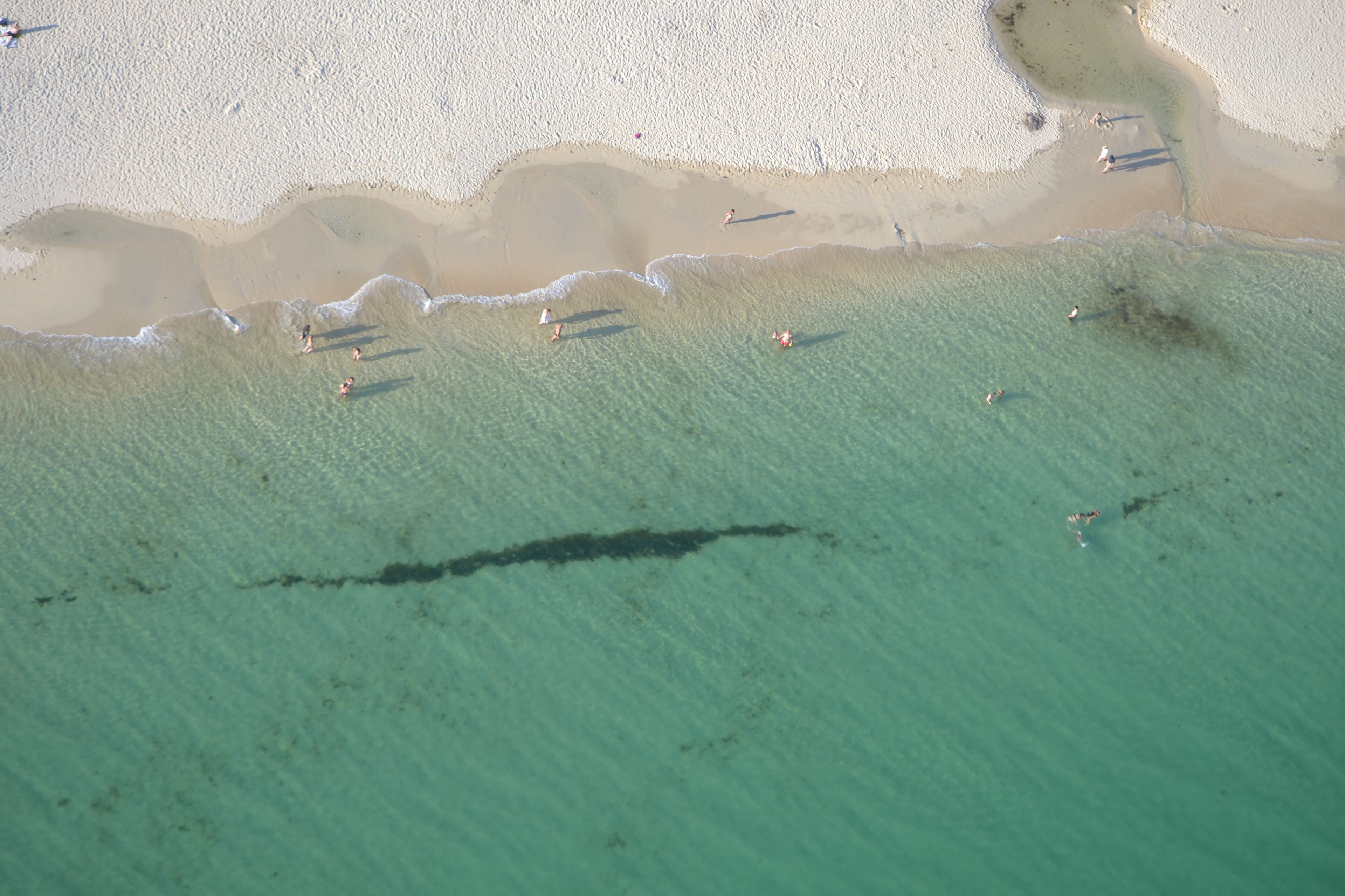 Une vue aérienne d’une plage aux eaux turquoise et limpides, avec un rivage sablonneux et quelques personnes dans l’eau.