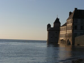 A historic stone building with a tower and a large windowed structure by the water. - In one of the most beautiful village of the Manche, functional garden level (Saint-Jean-Le-Thomas)