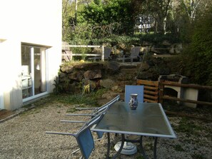 An outdoor patio with a table and chairs, a vase, and a garden area with rocks and plants. - In one of the most beautiful village of the Manche, functional garden level (Saint-Jean-Le-Thomas)