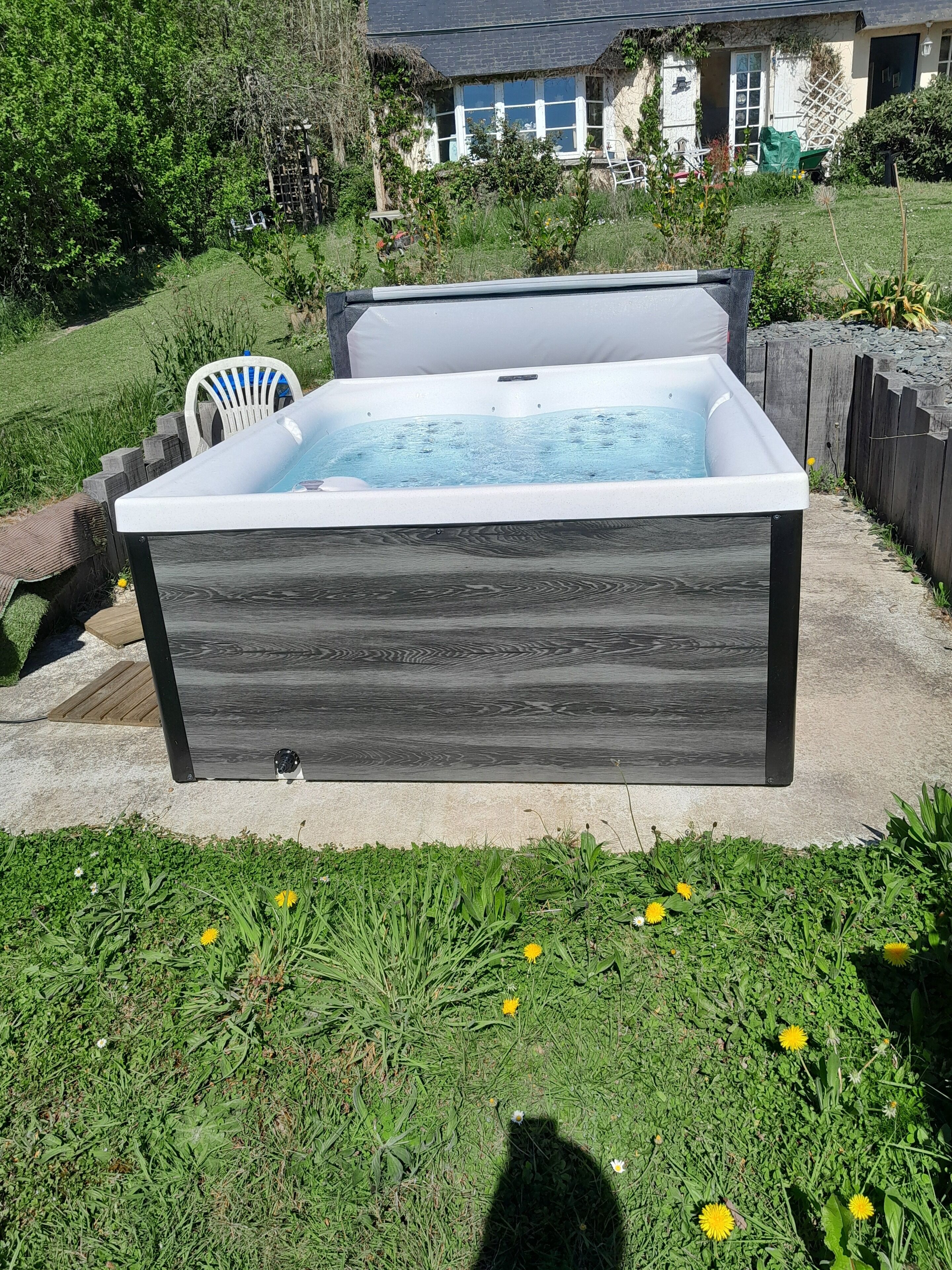 Un bain à remous rectangulaire, doté d’une structure extérieure en bois foncé, installé sur une terrasse en béton entourée de pelouse et de pissenlits.