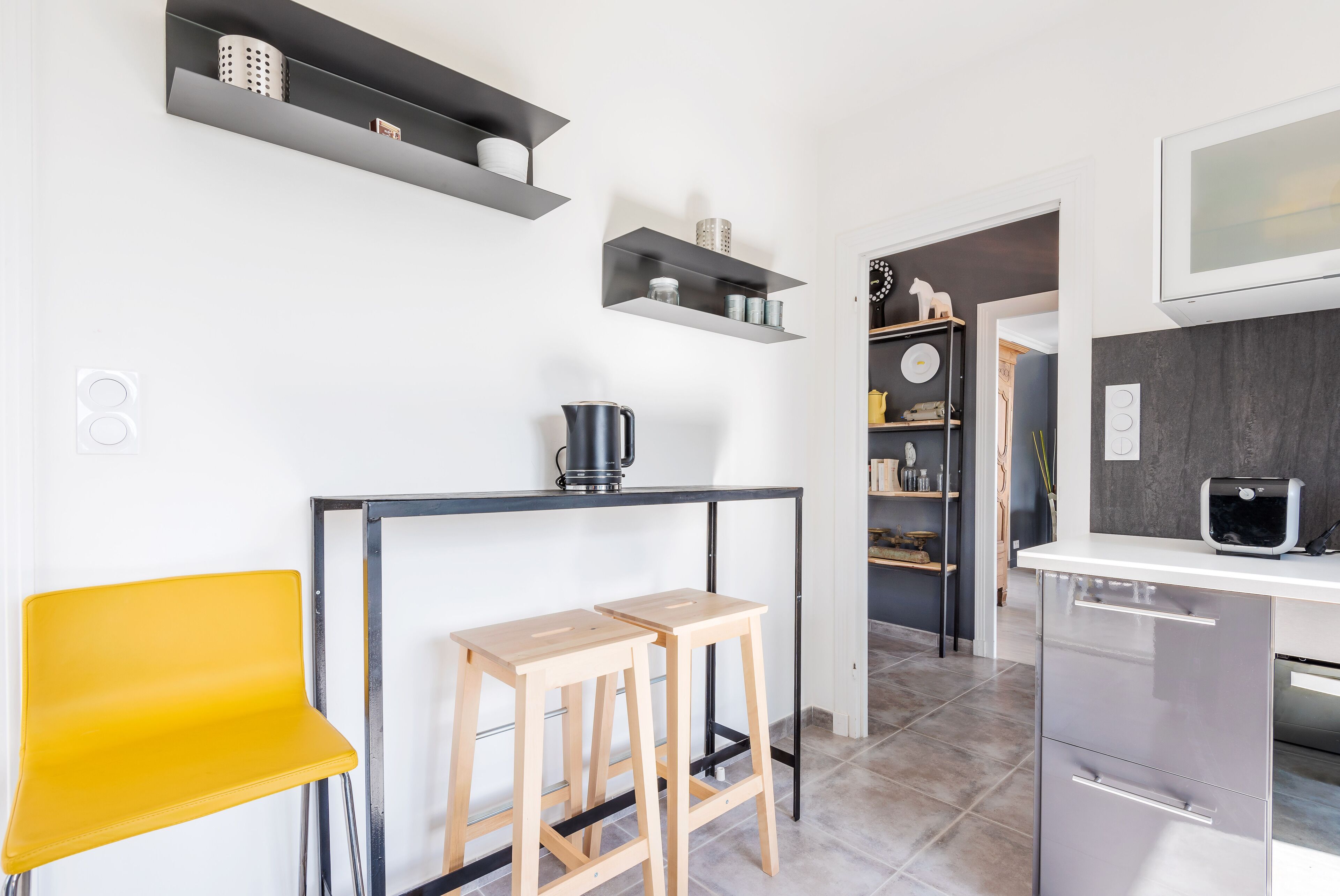 Una cocina moderna con encimera negra, dos taburetes de madera, una silla amarilla y estanterías empotradas.