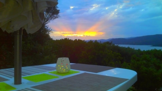 A table with a white tablecloth and a decorative object on it, overlooking a scenic view of a sunset over a body of water and hills.