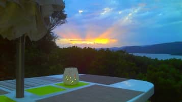 Une table recouverte d’une nappe blanche, agrémentée d’un objet décoratif, d’où l’on peut admirer un magnifique paysage : un coucher de soleil sur une étendue d’eau et des collines.