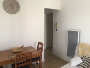 Une pièce comprenant une table à manger en bois, des chaises, un saladier en bois, une horloge murale, un lit avec un oreiller à rayures et un radiateur.