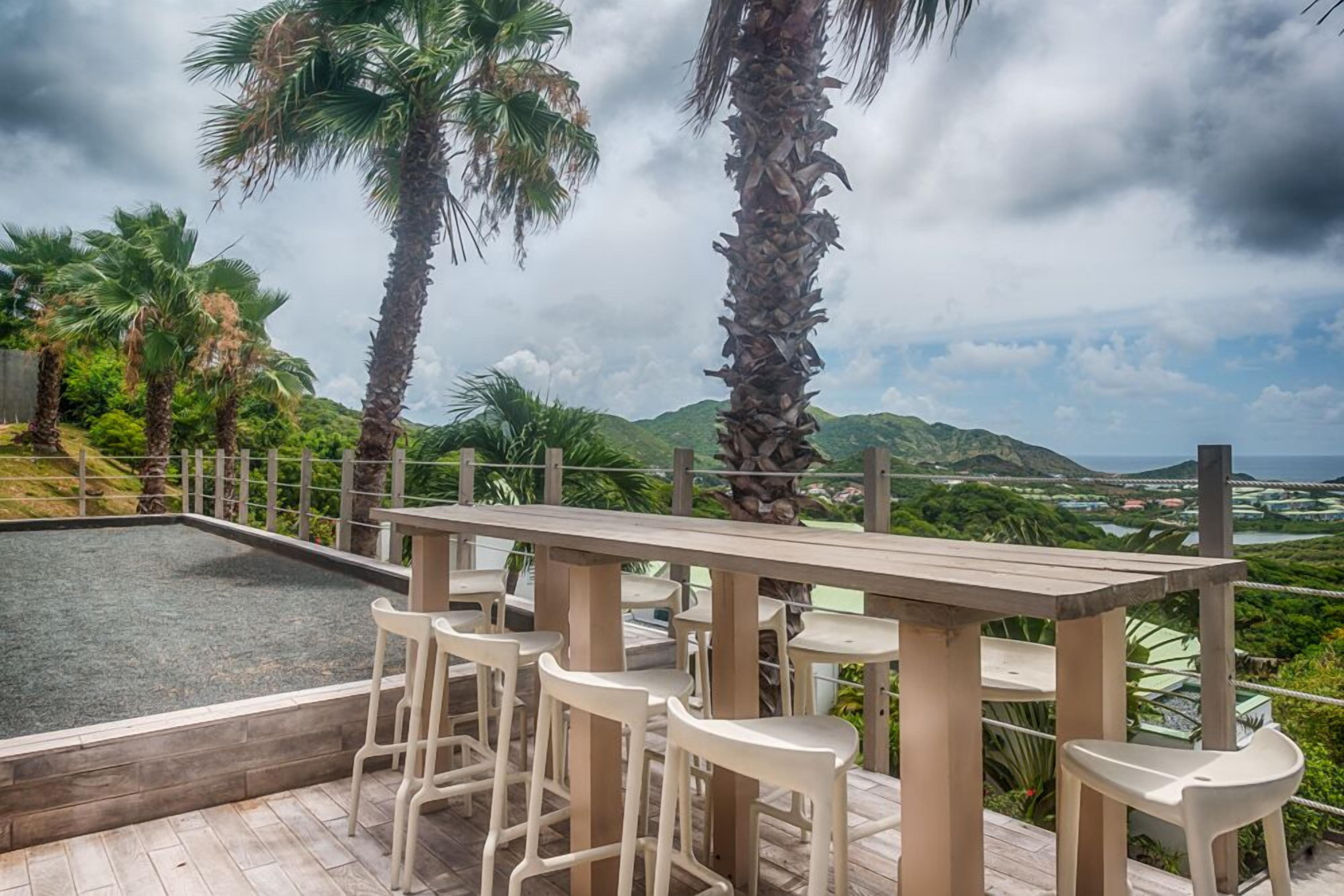 A poolside area with a wooden table and white plastic chairs, surrounded by palm trees and a view of green hills.