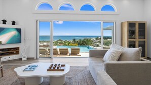Ett modernt vardagsrum med en stor TV, ett soffbord och en soffa. Det finns en skjutdörr i glas som leder ut till en balkong med en pool och utsikt över havet.