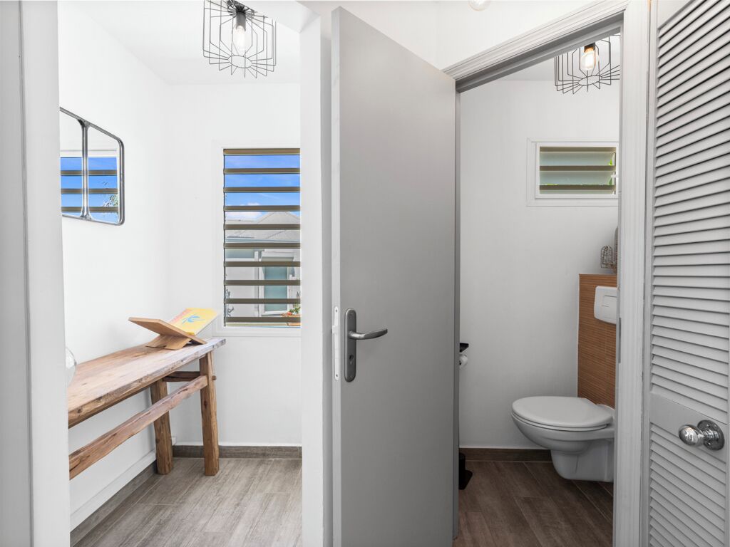 Une petite salle de bain comprenant des toilettes, une fenêtre avec des stores, un banc en bois et une porte dotée d’une poignée.