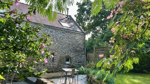 A stone building with a tiled patio, a small table and chairs, and a garden with various plants and flowers. - Country house heated pool 15 minutes from the Gulf of Morbihan (Sulniac)