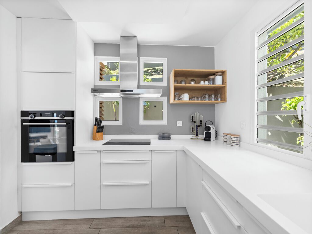 A modern kitchen with white cabinetry, a built-in oven, a stainless steel range hood, and a window with a view of greenery.