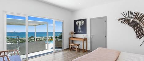 Une chambre dotée d’un grand lit, d’un bureau, d’une chaise et offrant une vue sur l’océan grâce à une porte-fenêtre coulissante.