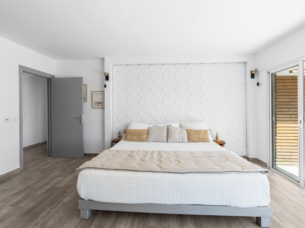 Ein modernes Schlafzimmer mit einem großen Bett, einer grauen Tür und einem Blick ins Freie durch eine Schiebetür.
