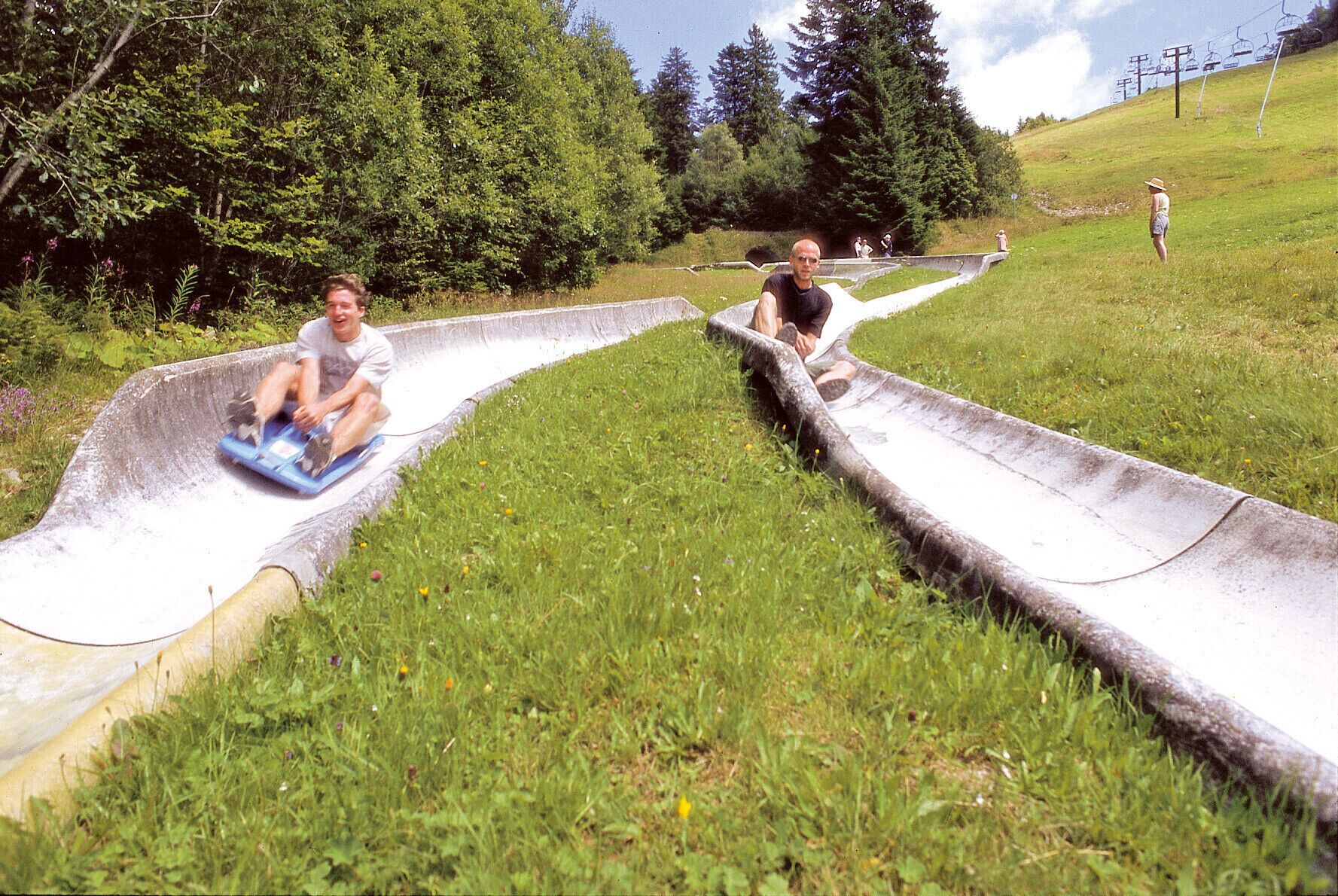 Twee personen glijden een betonnen glijbaan af in een grasrijk gebied, met bomen en een skilift op de achtergrond.
