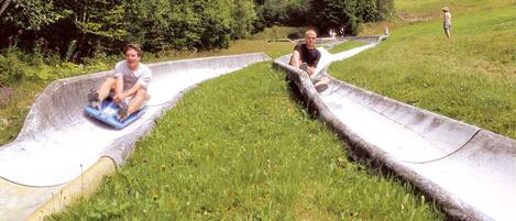 Twee personen glijden een betonnen glijbaan af in een grasrijk gebied, met bomen en een skilift op de achtergrond.