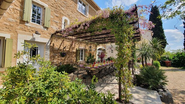 A stone building with a covered outdoor dining area, surrounded by greenery and a pergola with flowering vines.